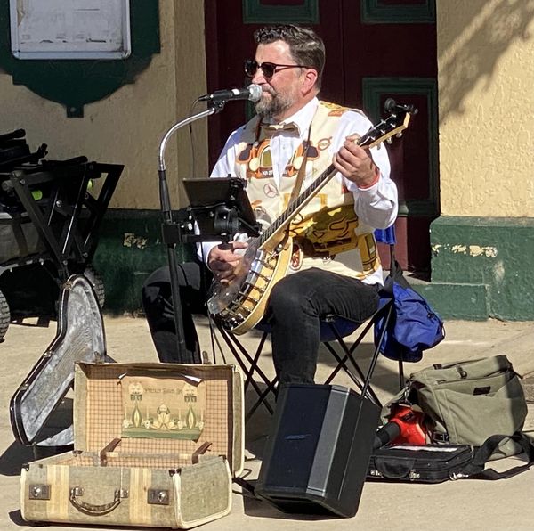 Photo of a man playing banjo with a suitcase open in front of him. He is wearing a vest with images of early motorcars on it.