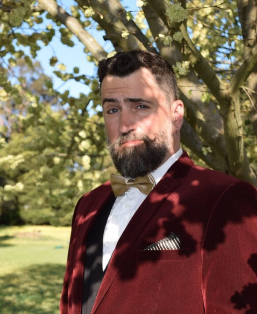 Image of Presley Davis Jr in a burgundy tuxedo standing under a eucalyptus tree with one raised eyebrow